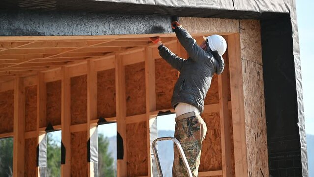 Male painter using paint roller, doing exterior paint work in a black color, standing on a ladder. Man worker building wooden frame house. Carpentry and construction concept.