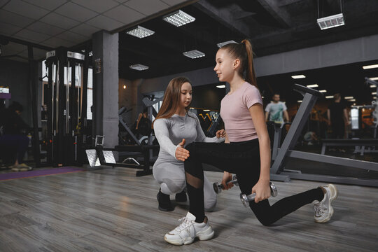 Teen girl doing lunges with weights, working out with personal trainer at the gym