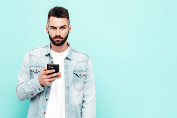 Handsome confident model.Sexy stylish man dressed in jacket and jeans. Fashion hipster male posing near blue wall in studio. Holding smartphone. Looking at cellphone screen. Using apps