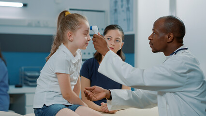 Physician using thermometer on kid to measure temperature in examination cabinet. General practitioner holding medical instrument to check if little child has fever. Health care