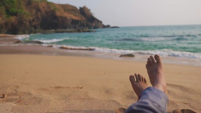 POV shot of legs of a man chilling on the tropical beach. Sandy feet of man in front of the blue tropical sea. Indian Man chilling at the Kakolem Beach in South Goa, India. Focus on the sandy legs.