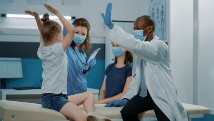 Medical team giving highfive to little child after successful examination, celebrating end of appointment. Medical assistant and pediatrician talking to cheerful girl at checkup visit.