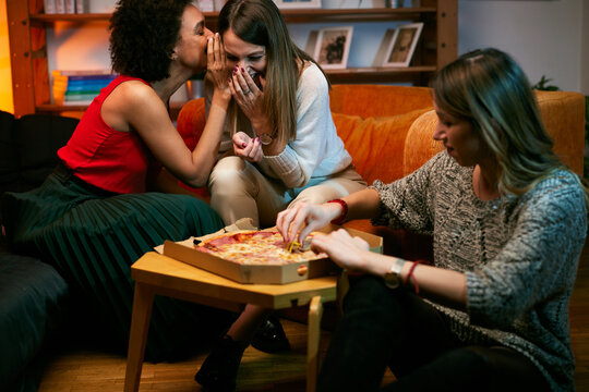 An african american girl telling secrets to a friend while other girl in front eating pizza.