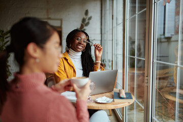 Two multicultural female students doing a school project on a laptop while drinking coffee in a cafeteria.