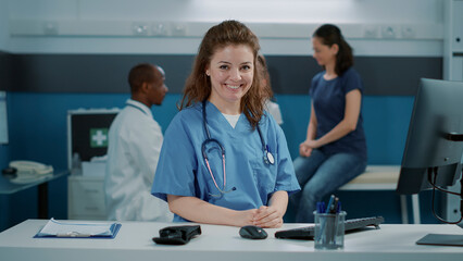 Portrait of woman nurse smiling and wearing uniform in office, sitting at desk. Medical assistant with stethoscope looking at camera and getting ready to help doctor at appointment.