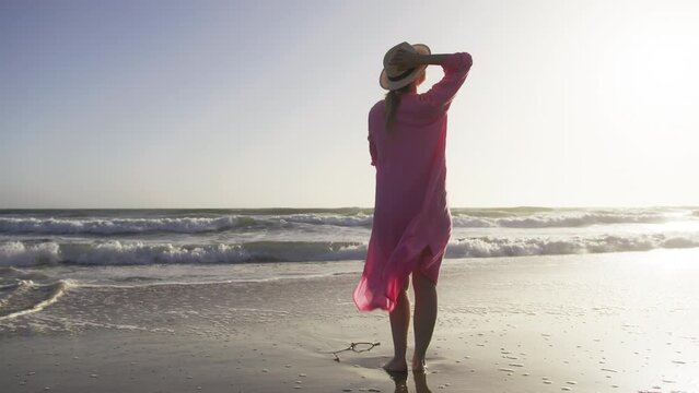 Young fit american woman smiling, embracing herself on cinematic sandy beach at blue stormy ocean with golden sunset light. Healthy outdoor leisure time by the sea. Woman in pink flattering dress RED