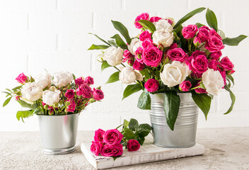 two fresh garden bouquets of roses in decorative buckets stand on a white wooden podium. grey concrete table. white brick wall-background.