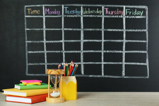 Hourglass and stationery on white wooden table near blackboard with drawn school timetable