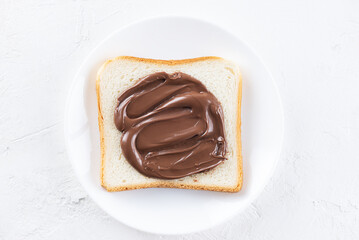 Toast bread spread with chocolate hazelnut spread in a plate on a white table, top view.