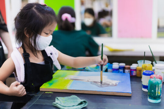 Portrait image of 5-6 years old childhood. Kindergarten child happy girl learning art watercolor in classroom. Kid wearing white cloth face mask to prevent spread virus and air pollution PM2.5.