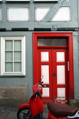 Moderner roter Motorroller vor einem alten grauen Fachwerkhaus mit schöner alter Haustür in Rot...
