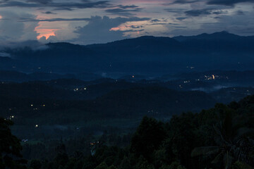 Indonesian tender landscape - colorful blue sky in evening with light pink clouds over hazy smoky blue mountains, green tropical valley, forest and villages with yellow lights on Bali, Munduk village.