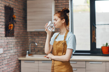cheerful woman drinking water in a brown apron kitchen interior home life