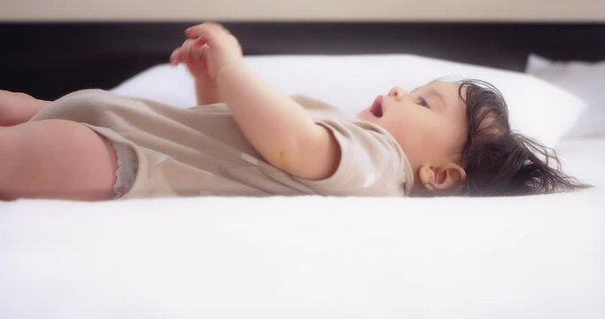 Cute girl in a beige bodysuit lies on her back on a white bed in the nursery,plays,laughing and chewing foot. Mom picks up her little baby - daughter in her arms.Concept of love, parenthood, childhood