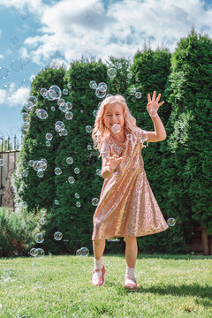 Happy smiling preschool fair-haired girl of 6-7 years old having light summer dress enjoying soap bubbles on meadow in city park on beautiful sunny summer day. Green nature background. Outdoor party