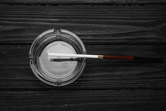 Cigarette in elegant holder and clean glass ashtray on black wooden table, top view