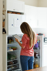 Fototapeta na wymiar Young woman standing next to an opened refrigerator door, holding a smartphone and ordering fresh fruit and vegetables online for home delivery. 