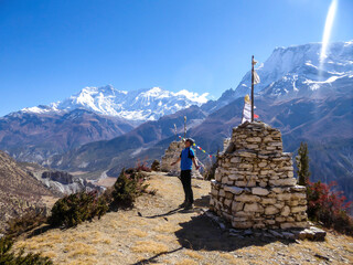 A young man standing next to a row of small stony stupas with Annapurna Chain as a backdrop, Himalayas, Nepal. High mountains covered with snow. Barren and dry land. Some prayer's flag next to it.