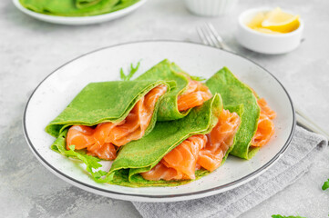 Green spinach crepes or pancakes with smoked salmon and soft cheese on a plate on the gray background. Copy space.