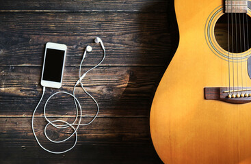 Naklejka premium Acoustic guitar on the wooden floor and the white ear phone smart phone and, soft light and shadow.