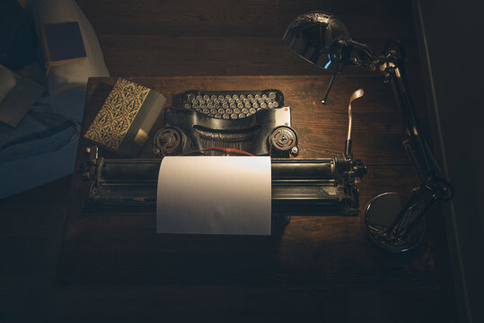An old classic typewriter in a vintage style studio with a wooden desk lit by a lamp, wit old books all around. A moody, dark atmosphere for a writer working at night.