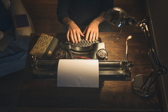Typing hands of a writer looking for inspiration to start a new novel on his vintage typewriter in a retro style studio: a wooden desk lit by a lamp, old books and a pair of glasses.