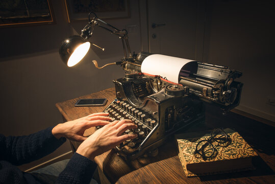 Typing hands of a writer looking for inspiration to start a new novel on his vintage typewriter in a retro style studio: a wooden desk lit by a lamp, old books and a pair of glasses.
