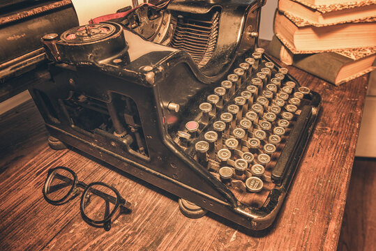 An old classic typewriter in a vintage style studio with a wooden desk lit by a lamp, wit old books all around. A moody, dark atmosphere for a writer working at night.