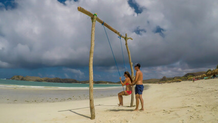 Fototapeta na wymiar A man pushing a girl on a swing placed on the seashore of Tanjung Aan Beach, Lombok, Indonesia. The swing has simple wood construction. Waves wash the pillars of it. In the back there are few boats.