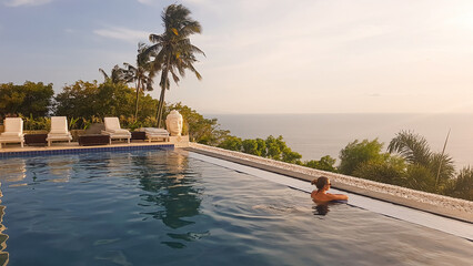 A young woman leaning at the edge of an infinity pool and looking at the sun setting into the sea. There is a big palm tree in front. Calmness and serenity. Luxury hotel in Lombok, Indonesia. © Chris