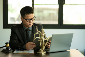 Middle aged Asian male lawyer working on laptop and legal contract documents in courtroom Judge's Hammer and Goddess Scales concept of legal counseling and services.