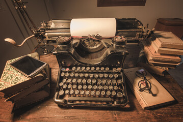 An old classic typewriter in a vintage style studio with a wooden desk lit by a lamp, wit old books all around. A moody, dark atmosphere for a writer working at night.