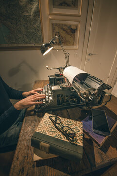 Typing hands of a writer looking for inspiration to start a new novel on his vintage typewriter in a retro style studio: a wooden desk lit by a lamp, old books and a pair of glasses.