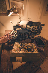 Typing hands of a writer looking for inspiration to start a new novel on his vintage typewriter in a retro style studio: a wooden desk lit by a lamp, an old book and a pair of glasses.