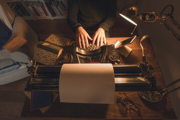 Typing hands of a writer looking for inspiration to start a new novel on his vintage typewriter in a retro style studio: a wooden desk lit by a lamp, old books and a pair of glasses.