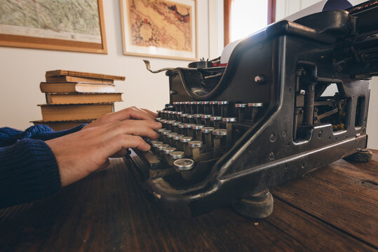 Typing hands of a writer looking for inspiration to start a new novel on his vintage typewriter in a retro style studio: a wooden desk lit by a lamp, old books and a pair of glasses.