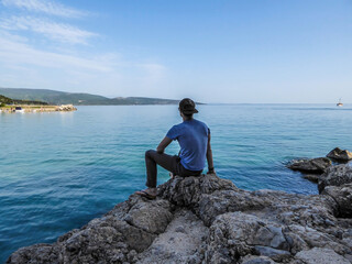 Fototapeta na wymiar Man is a full cap sits at the stony shore of the Mediterranean Sea and looks at the calm surface of the water. He is calms and peaceful. Water has many shades of blue. In the back there are islands