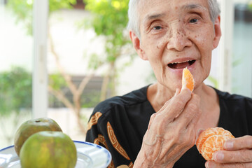 Happy healthy senior woman eating tangerine deliciously,asian old elderly opening her mouth wide to...