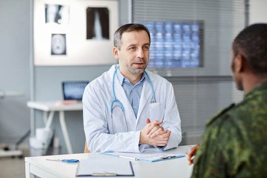 Mature Caucasian physician sitting at desk in front of unrecognizable Black soldier speaking about tests needed for health checkup