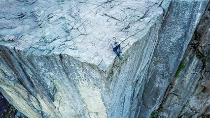 Areal shot of a young man sitting at the edge of Preikestolen. First morning sunbeams reach the cliff formation. Massive rocks surrounding the man. Dangerous place for a hike. Beauty of the nature