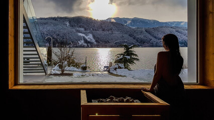 Girl standing next to a window in an alpine sauna with the view on the lake and the mountains....