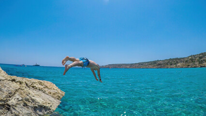 A young athletic-build man jumping into the sea in Cape Greco, Cyprus. Water is crystal clear, shimmering with many shades of blue. Jump of joy. Man is wearing blue swimsuit.