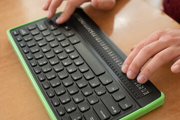 A blind man uses a computer with a Braille display and a computer keyboard. Inclusive device. - obrazy, fototapety, plakaty