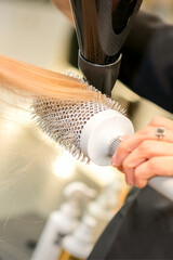 Drying straight blond hair with black hairdryer and white round brush in hairdresser salon, close up