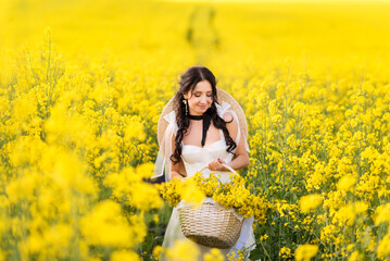 Naklejka na ściany i meble beautiful woman collects yellow rapeseed flowers in a large basket. A girl in a white dress and a big hat