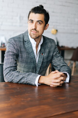 Vertical portrait of confident stylish business man in fashion suit sitting at desk in modern office room, looking at camera. Front view of serious bearded businessman posing at workplace.