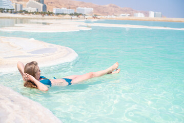 Slim woman in green bikini swim and lies in blue water of Dead Sea. Very salty water push out. Treatment and recreation in Israel, Dead Sea, salt and mud are good for health.