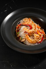 Funchoza with shrimp and vegetables on a black background, top view, copy space. Glass noodles, Asian food.