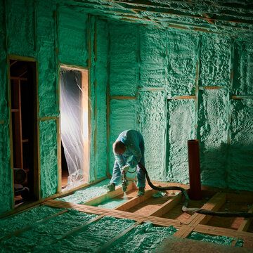 Male builder insulating wooden frame house. Man worker spraying polyurethane foam on floor inside of future cottage, using plural component gun. Construction and insulation concept.