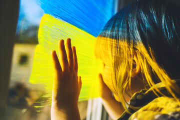 Little child with hands on yellow-blue flag image of Ukraine on window. Support Ukrainians. Concept, emotions. Color symbol image of flag of Ukraine on glass with paints.